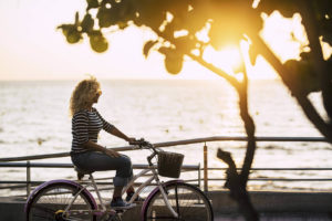 Photo of a woman riding a bike beside of a body of water during sunset. Learn to find peace with your unresolved trauma by starting trauma therapy in Marin County, CA.
