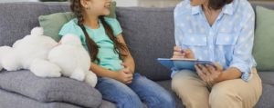 Photo of a young girl sitting on a couch smiling while talking to a child therapist. This photo represents how therapy for children in Marin County, CA can help your child manage their emotions and behaviors in a healthy way.