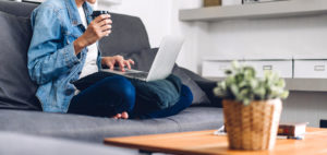 Photo of a woman sitting on her couch using a laptop and drinking coffee. This photo represents how easy it can be to get online therapy in San Francisco, CA. Learn how online therapy in California can help you with your anxiety and depression. 