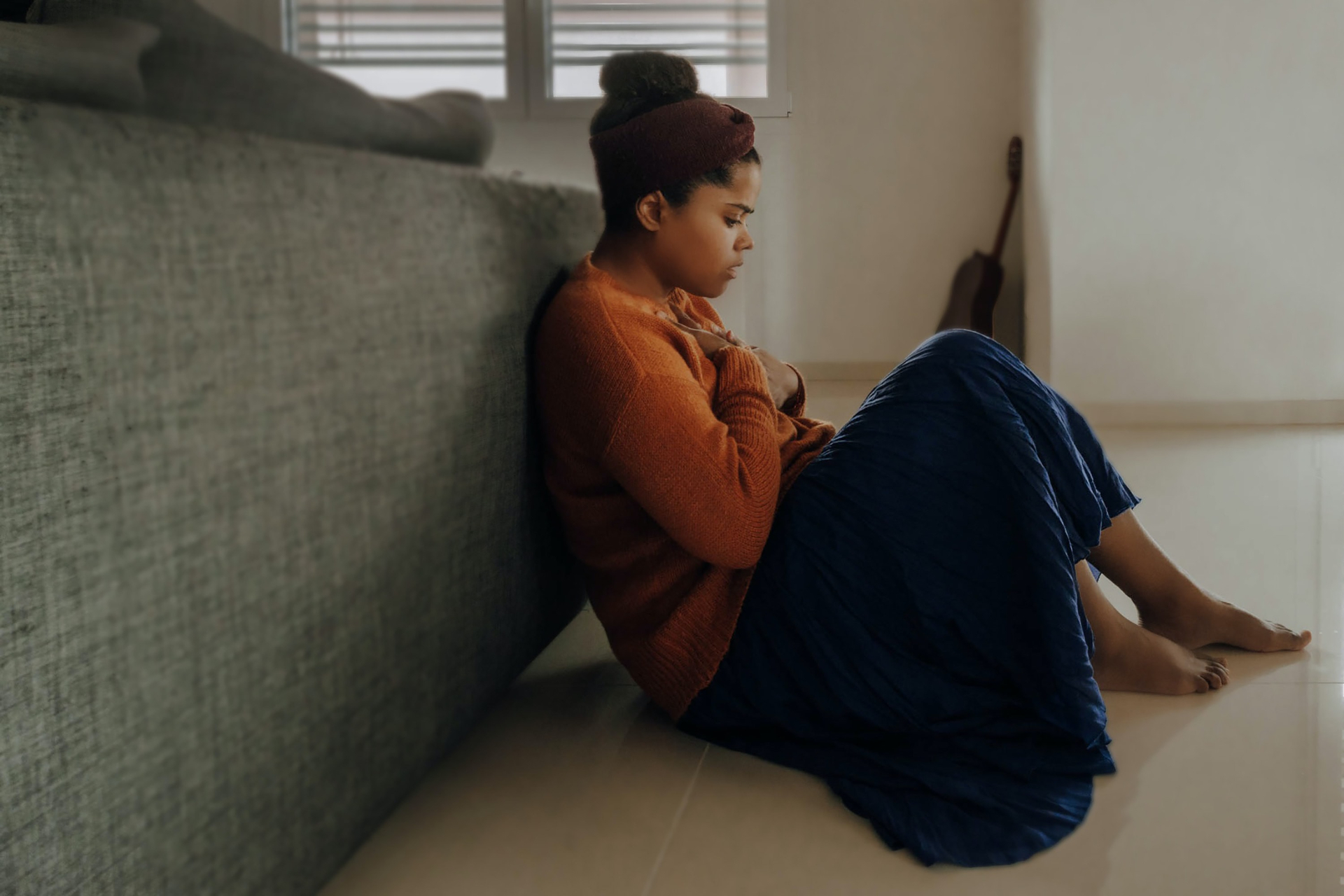 Photo of an African American woman sitting on the floor with her hands clutching her chest. This photo represents how postpartum symptoms can affect your daily life and how postpartum therapy in San Francisco, CA can help you begin to cope.
