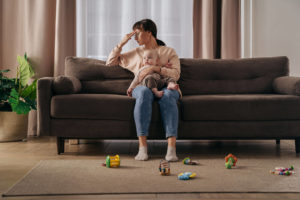 Photo of a mother sitting on a couch with her baby on her lap holding her head. This photo represents how motherhood can bring emotional challenges to being a parent. Learn how counseling for mothers in San Francisco, CA can help you manage.